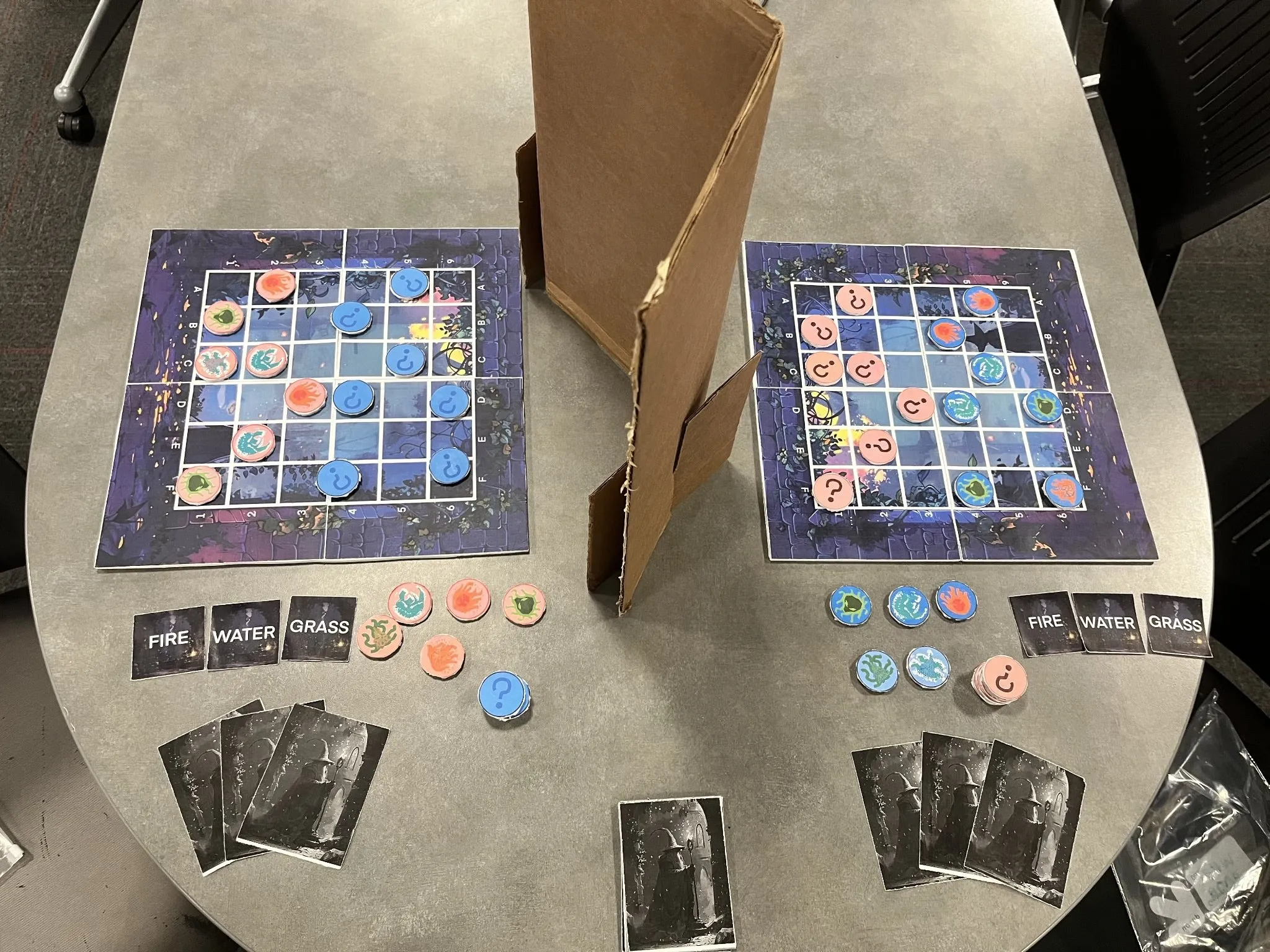 Two Spell Showdown boards on a table with a cardboard divider between them, each board holding one team's minion tokens and question-mark markers for the other team